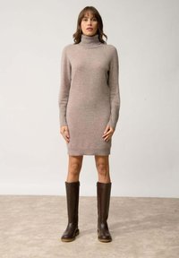 Abito a maniche lunghe con collo alto in tessuto a maglia beige chiaro, con una forma aderente e orlo a coste. Indossato con stivali alti in pelle marrone.