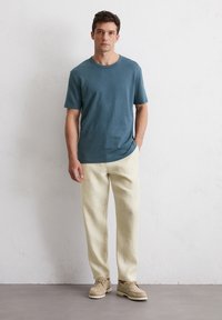 Blauwe T-shirt met korte mouwen, relaxte pasvorm; beige broek, rechte pijpen met zijzakken. Licht schoeisel, natuurlijke tinten, gestructureerd oppervlak.