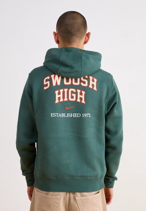 Osoba w ciemnozielonej bluzie z kapturem założonym na głowę, tekst "Swoosh High" i logo Nike z "Established 1972" na plecach, widoczne beżowe spodnie.