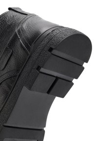 Zapato de cuero negro con una superficie texturizada, que cuenta con una suela de goma gruesa y con patrón, y ranuras de tracción definidas en la parte inferior.