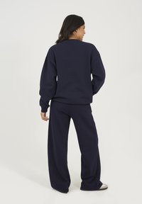 Donna con il retro rivolto, che indossa una felpa blu navy e pantaloni larghi, con scarpe bianche e una fascia nera sui capelli, su uno sfondo neutro.