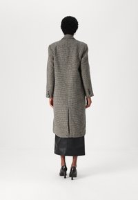 Abrigo largo de pata de gallo gris con un ajuste estructurado, abertura en la parte trasera y puños de tres botones, combinado con una falda de cuero negra y zapatos de tacón.