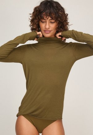 Vrouw met krullend haar die een olijfgroene coltrui met lange mouwen afstelt, draagt bijpassend ondergoed, tegen een effen lichte achtergrond.