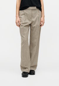 Άτομο που φοράει μπεζ καρό παντελόνι με φαρδιά πόδια και μαύρα στιλπνά μοκασίνια, στέκεται μπροστά σε απλό φωτεινό φόντο.