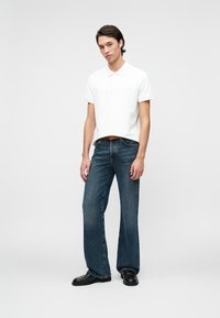 Bărbat în picioare, îmbrăcat cu un polo alb cu mânecă scurtă, blugi dilatați de culoare albastră închis, curea maro și pantofi negri de birou, pe un fundal simplu și deschis.