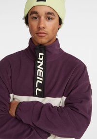 Pullover in pile viola e bianco con collo alto e zip a metà, caratterizzato da un pannello nero con il logo "O'Neill." Texture morbida.