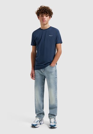 Teenager-Junge, der ein marineblaues T-Shirt, hellblaue Jeans und weiße Turnschuhe trägt, vor einem schlichten grauen Hintergrund steht.
