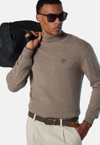 Maglione a collo alto marrone realizzato in materiale morbido, con un piccolo dettaglio logo. Abbinato a una cintura intrecciata verde e pantaloni di colore chiaro.