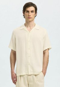 Camicia a maniche corte, di colore beige chiaro, realizzata in lino, con chiusura a bottoni, colletto classico e vestibilità rilassata, abbinata a pantaloni beige coordinati.