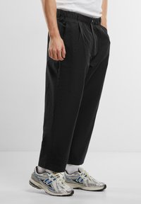 Pantaloni negri dintr-un material ușor, cu talie elastică și două buzunare frontale, cu un croi relaxat și plii.