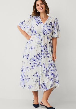 Kvinde smiler, iført en hvid midikjole med blå blomsterprint, korte puffede ærmer, knapper foran og sorte lave hælede slingback-sko.