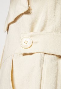 Beige textilní látka zblízka s krémovým knoflíkem se čtyřmi dírkami a raženým textem. Knoflík je upevněn na složené části límce.