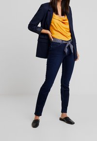 Jeans ajustados de color azul oscuro combinados con una blusa amarilla de satén con cuello drapeado y una chaqueta azul marino. Los jeans tienen un cinturón con estampado y zapatos negros de tipo slip-on.