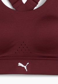 Reggiseno sportivo bordeaux con spalline incrociate, caratterizzato da perforazioni decorative e un logo Puma bianco in basso a destra.