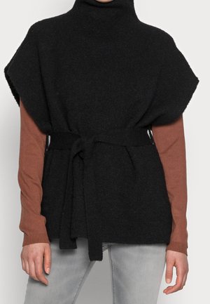 Czarna dzianinowa kamizelka bez rękawów z wysokim kołnierzem i paskiem zawiązanym w talii, noszona na brązowej bluzce z długim rękawem oraz jasnoszarych spodniach.