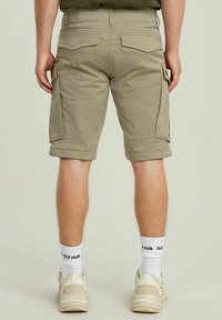 Pantaloni cargo beige con doppie tasche laterali, orli arrotolati e tasche posteriori. Indossati con calze bianche e sneaker chiare.