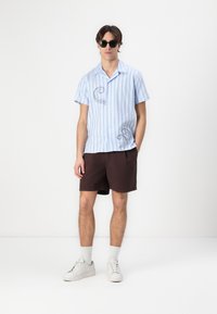 Camicia a maniche corte a righe blu con design di polpo, abbinata a pantaloni corti marroni e sneaker bianche, con calze bianche fino alla caviglia.