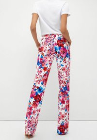 Vrouw in een witte aansluitende t-shirt en hoge taille, brede broek met een levendig patroon van roze, rode en blauwe bloemen, staand met handen in de zakken.