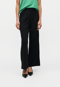 Γυναίκα που φοράει μαύρο παντελόνι φαρδιάς γραμμής, πράσινο τοπ και μαύρες μυτερές γόβες, στέκεται μπροστά σε απλό λευκό φόντο.