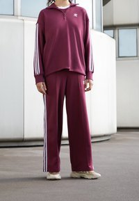 Conjunto deportivo burdeos que incluye una camiseta de Manga Larga con cremallera y franjas blancas y pantalones a juego, ambos con un pequeño logo en el pecho y el muslo.