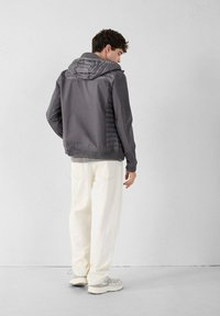 Grijze jas met een opgezette capuchon, gestructureerde zijpanelen, geribbelde boorden en zoom, gecombineerd met witte broek en grijze sneakers. Achteraanzicht.