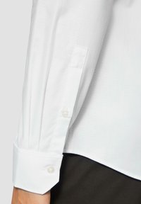 Chemise blanche à manches longues avec une texture en chevrons, poignets classiques avec des boutons en nacre et une coupe ajustée visible au niveau de l'ourlet.