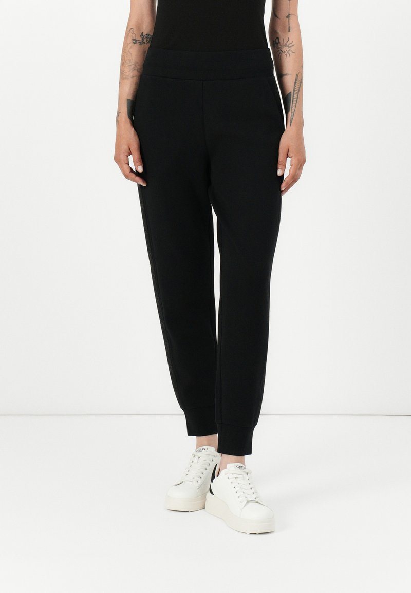 Svarte joggebukser laget av mykt stoff, med bred elastisk midjebånd, ribbede mansjetter og sidelommer. Kombinert med hvite joggesko.