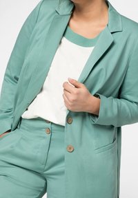 Blazer verde menta con rever a punta, bottoni singoli e pantaloni coordinati. Sotto, una maglietta color crema. Tessuto liscio, vestibilità su misura.