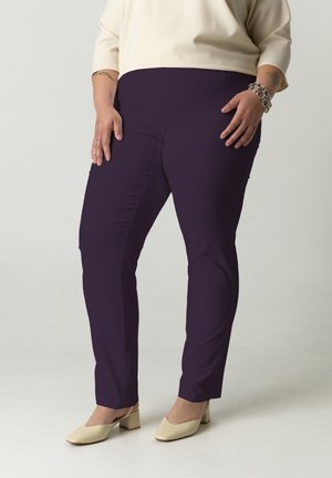 Person, die dunkelviolette, eng anliegende Hosen, kremfarbene Schuhe mit niedrigen Absätzen, ein cremefarbenes Oberteil und ein silbernes Kettenarmband trägt, steht vor einem einfarbigen Hintergrund.