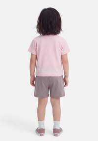 Παιδί που στέκεται με την πλάτη γυρισμένη, φορώντας ανοιχτό ροζ κοντομάνικο πουκάμισο, γκρι σορτς, λευκές κάλτσες και ροζ αθλητικά παπούτσια σε λευκό φόντο.