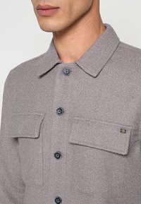 Grijs gestructureerd shirt met kraag, voorzien van twee borstzakken en donkere knopen. Opvallende details zijn onder andere een klein logo op de zak.