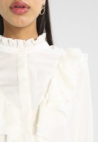 Bílá blůza s volánkovými detaily po límci a ramenou, s hladkou texturou a knoflíkovým zapínáním u krku.