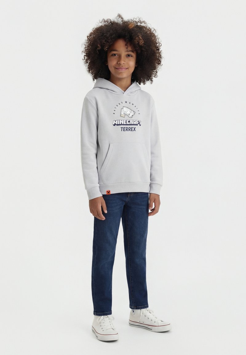 Enfant aux cheveux bouclés portant un sweat à capuche Minecraft Terrex gris clair, un jean bleu foncé et des baskets blanches montantes, debout et souriant.