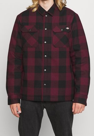 Mężczyzna w bordowej i czarnej koszulowej kurtce w kratę z dwoma kieszeniami na piersiach oraz czarnych dżinsach.