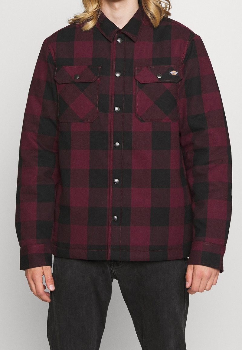 Homme portant une veste-chemise à carreaux bordeaux et noir avec deux poches poitrine et un jean noir.
