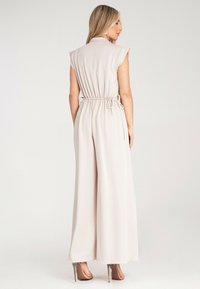 Beżowy, bezrękawnikowy kombinezon z kołnierzykiem, z wcięciem w talii i szerokimi nogawkami. Gładki materiał o delikatnym zwisie, zestawiony z beżowymi szpilkami.