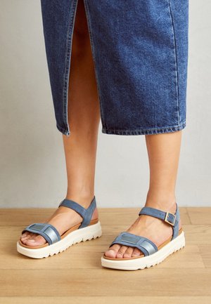 Fødder iført blå remsandaler med hvide såler, vist med en knælang blå denimnederdel med frontslids.