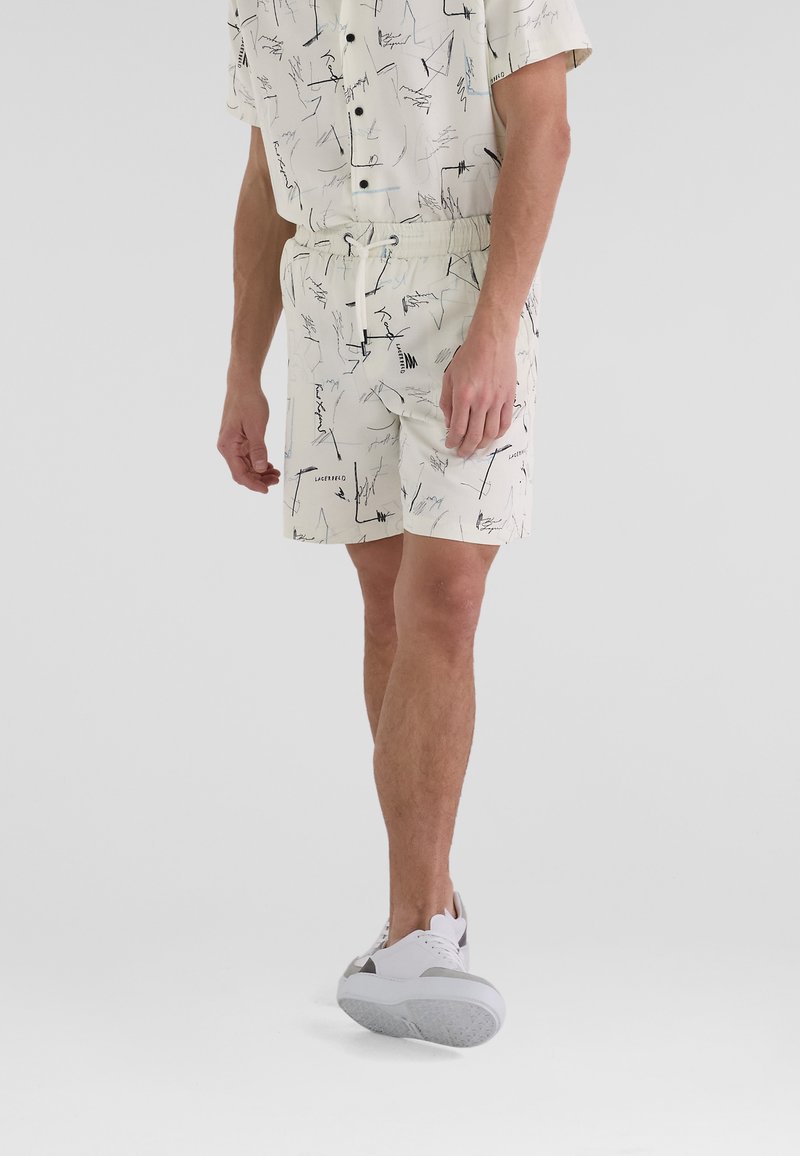 Hellebeige gemusterte Shorts mit elastischem Bund, die mit schwarzen und blauen abstrakten Designs versehen sind, kombiniert mit weißen Sneakers.