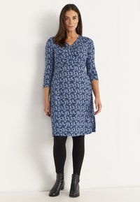 Frau steht in einem blauen, floralen knielangen Kleid mit dreiviertel Ärmeln, schwarzen Leggings und schwarzen Stiefeletten vor einem schlichten Hintergrund.