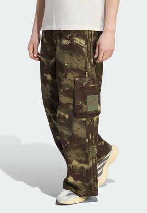 Camouflage cargo pants with large side pocket featuring Adidas logo, worn with white and beige sneakers and a plain white T-shirt.