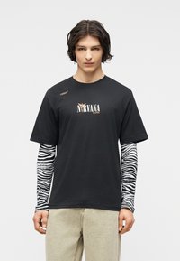 Άνδρα ς με κυματιστά σκούρα μαλλιά, φοράει μαύρο T-shirt Nirvana πάνω από μακριά μανίκια με ζεβρέ ρίγες και μπεζ παντελόνι, στέκεται μπροστά σε απλό φόντο.