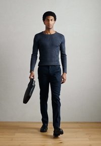 Marinblå ribbad långärmad tröja kombinerad med mörka byxor, håller en svart portfölj, står på trägolv framför en enfärgad vägg.