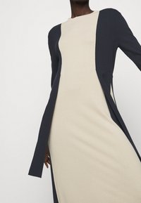Gebreide jurk met een beige lichaam en donkerblauwe accenten, lange mouwen en zijsplitten, die een gestructureerd maar vloeiend ontwerp benadrukken.