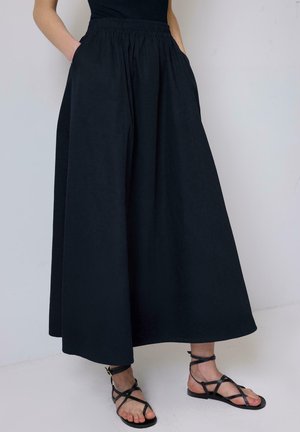 Black, flowy maxi skirt made of lightweight fabric, featuring an elastic waistband and side pockets. Paired with simple black strappy sandals.