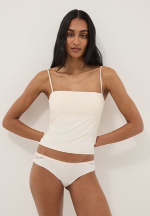 Frau mit langen dunklen Haaren, die ein cremefarbenes Camisole-Top und passende Unterwäsche mit Spitzenbesatz trägt, steht vor einer einfarbigen Wand.