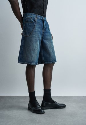 Άτομο που φοράει μπλε τζιν σορτς μεσαίου μήκους με αλυσίδα ως αξεσουάρ, μαύρες κάλτσες και μαύρα δερμάτινα loafers σε γκρι πάτωμα.