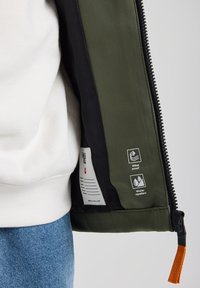 Olive green jacket interior with black lining, featuring windproof and water-repellent icons. Zipper and orange drawstring detail visible.