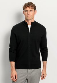 Schwarzer Reißverschluss-Pullover mit hohem Kragen, langen Ärmeln und taillierter Passform, aus einem glatten, strukturierten Material.