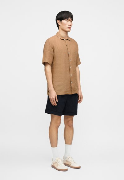 Chemise en lin marron à manches courtes avec un devant boutonné, associée à un short bleu marine et des baskets claires. Le mannequin se tient devant un fond uni.