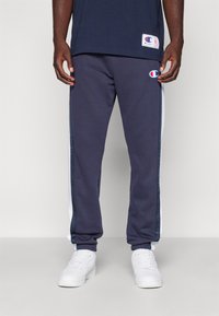 Pantalones jogger para hombre en color azul marino con franjas laterales blancas, puños elásticos y cintura elástica. La tela parece suave y presenta un pequeño logo en el muslo izquierdo.